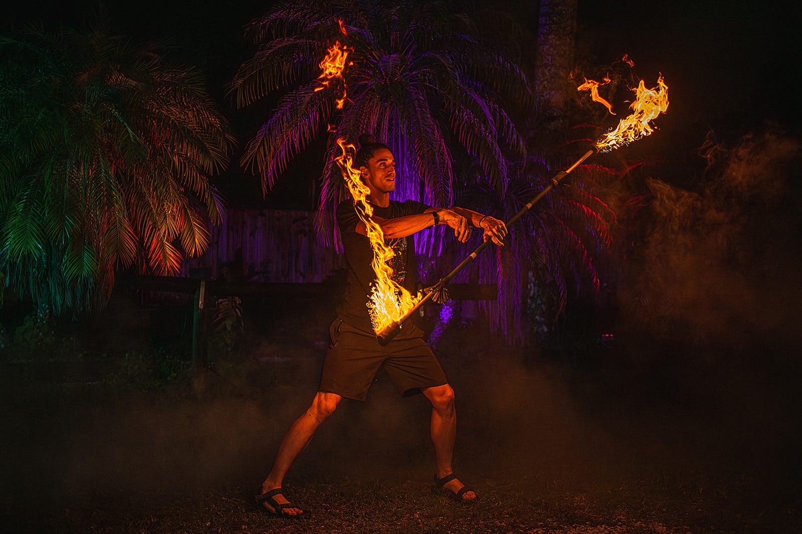 Jake in a wide stance spinning a fire staff lit at both ends, purple-lit palms behind him