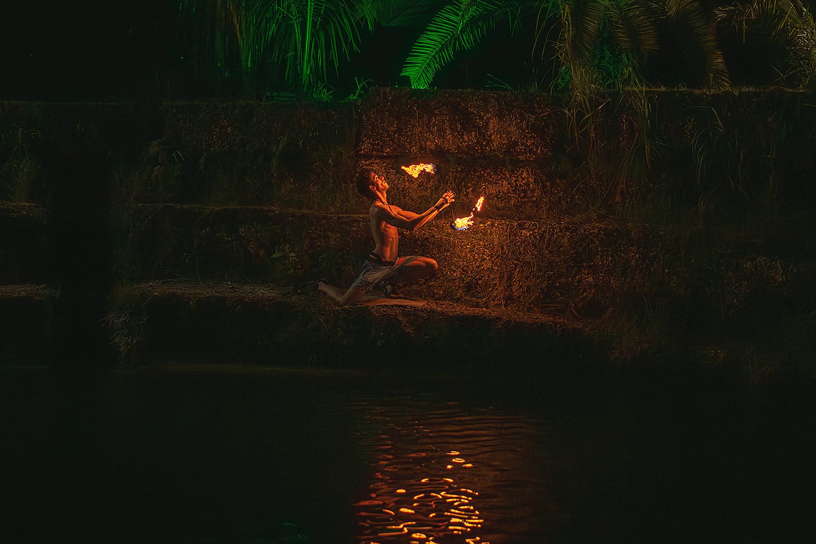 Jake kneeling on a stone ledge at night, holding a lit fire prop up in both hands, the flame reflected in the water below