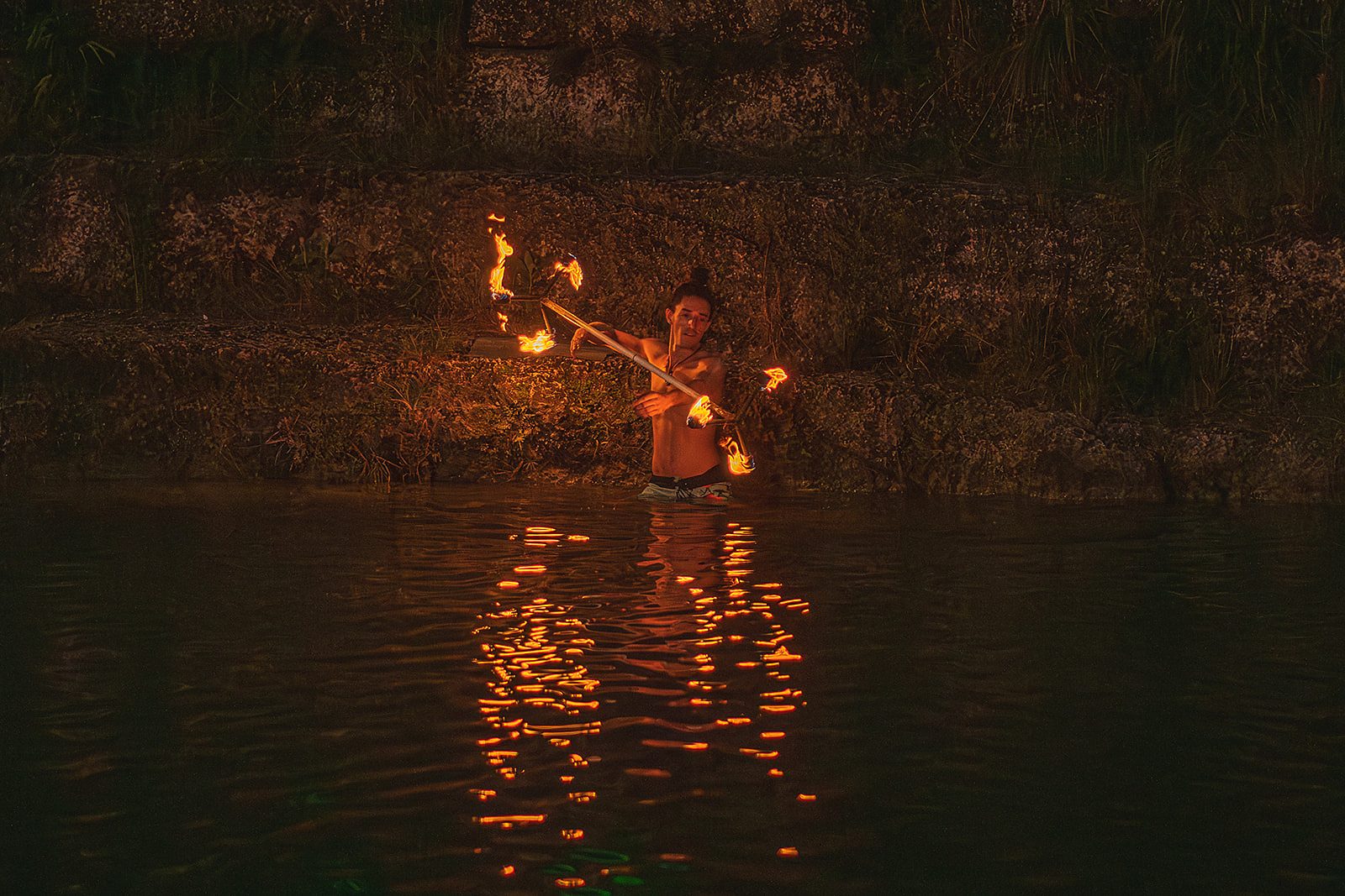 Fire staff spinning above dark water, its reflection rippling toward the camera