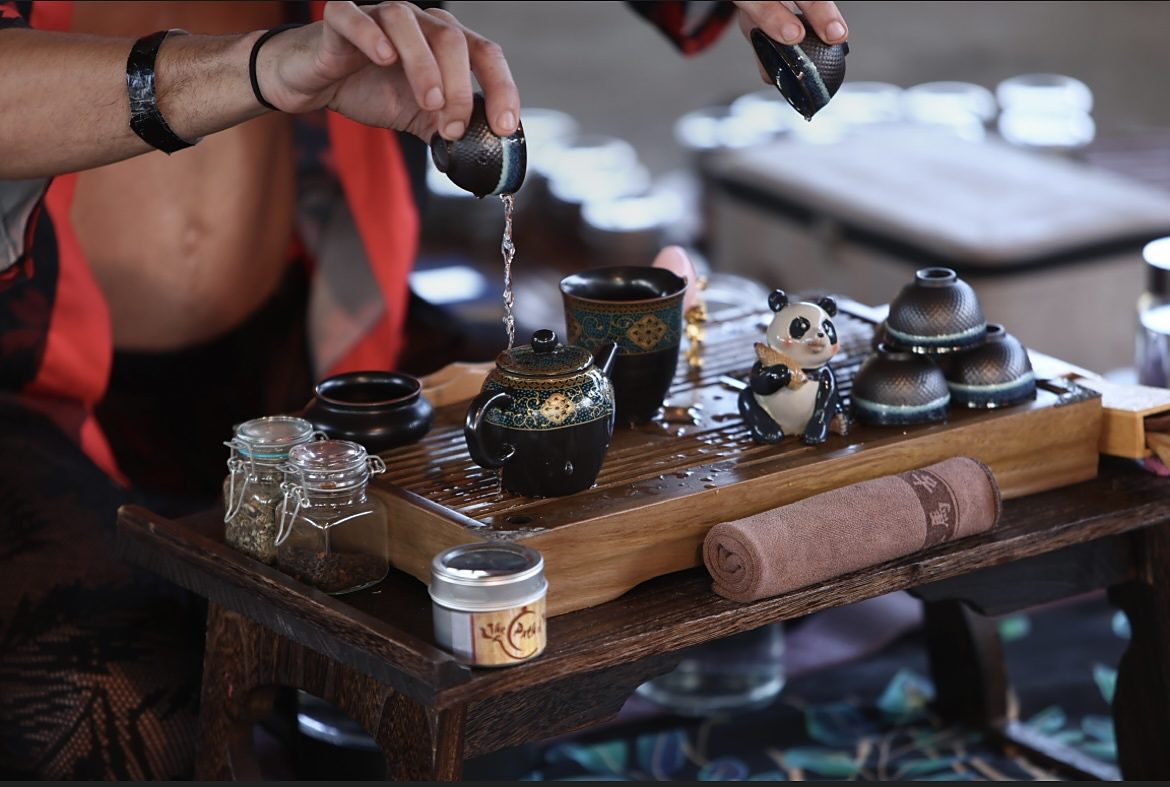 Jake's hands pouring tea over a wooden tray set with a patterned teapot, dark cups, jars of loose leaf and a small ceramic panda